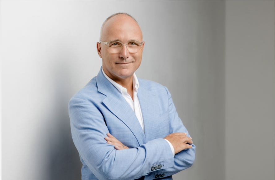 Smiling bald man wearing glasses and a light blue suit jacket with arms crossed, standing against a light gray background.