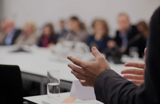 Speaker gesturing with hands during a meeting while audience listens around a conference table.