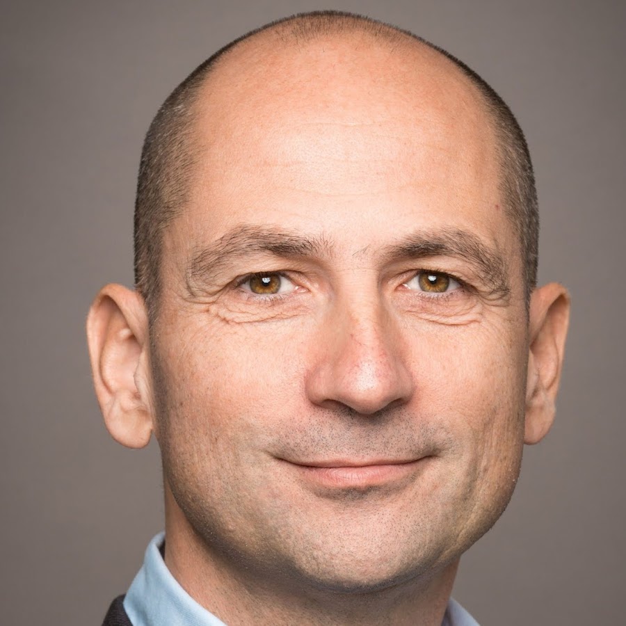 Close-up portrait of a middle-aged man with a shaved head, light brown eyes, and a slight smile, wearing a light blue shirt and dark jacket with a neutral gray background.
