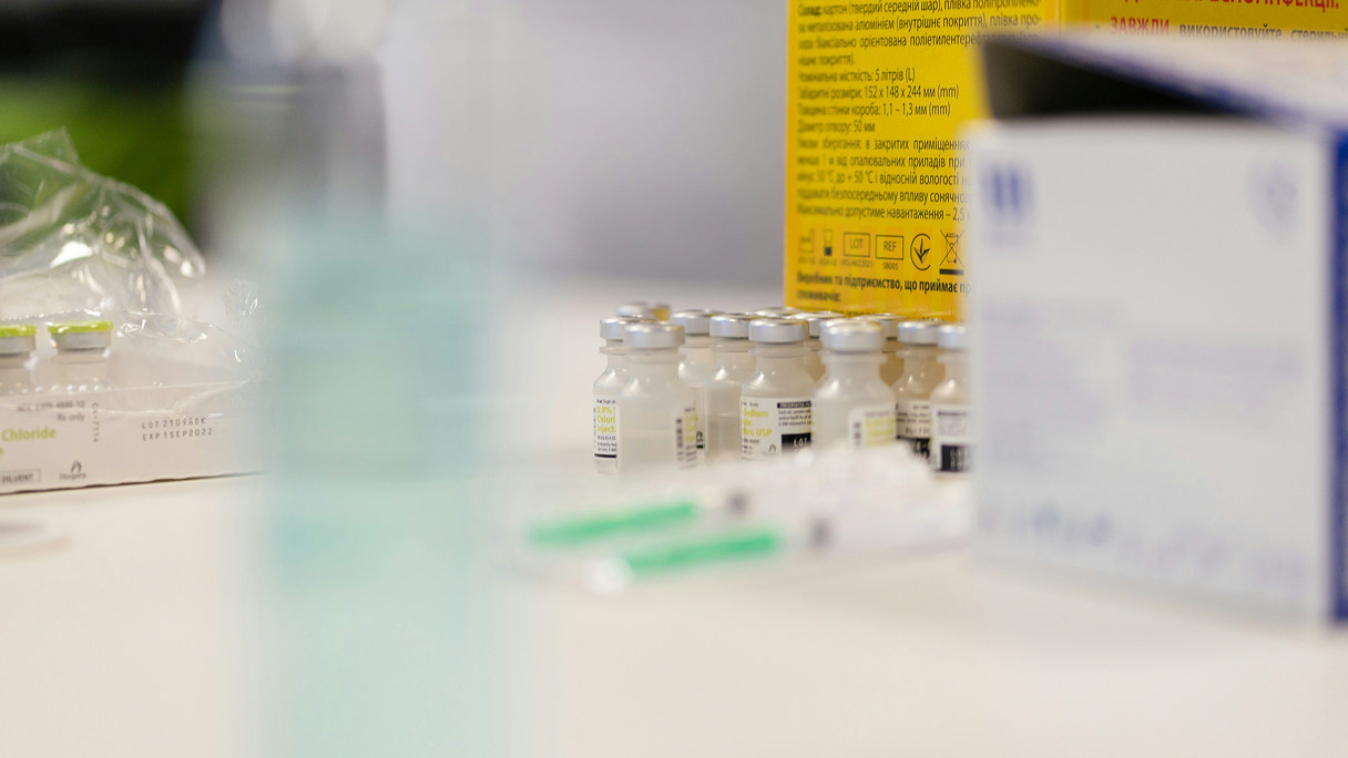 Small clear vaccine vials with silver caps grouped together on a table surrounded by medical packaging and syringes.