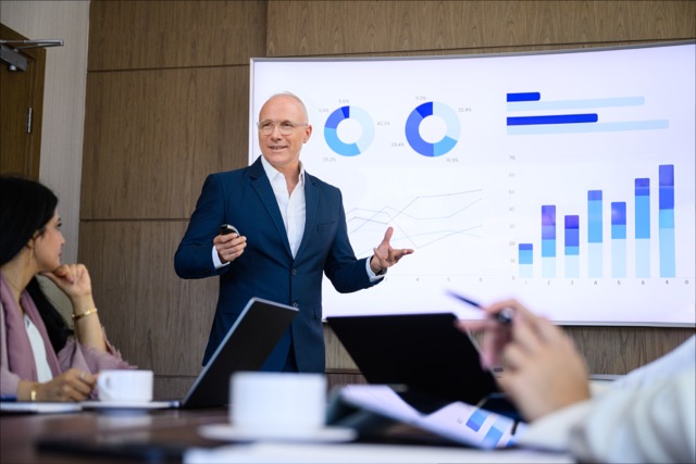 A man in a suit giving a presentation in a conference room with charts displayed on a large screen, while colleagues listen attentively.